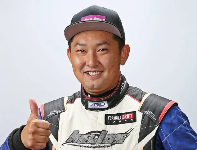 FORMULA DRIFT JAPANのレーシングスーツ姿で、親指を立てて笑う飯塚一哉